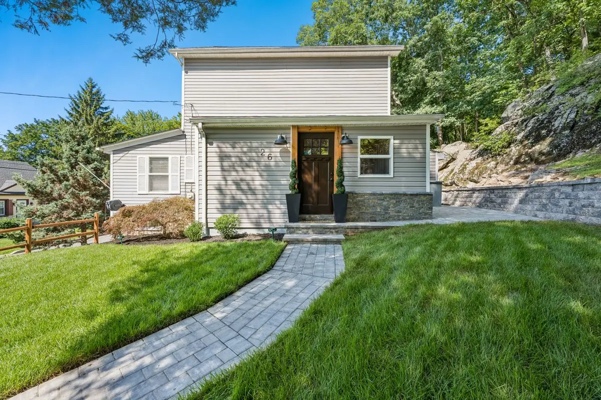 Daytime front view of 26 Oak Street under a blue sky — the gray Colonial with a stone paver walkway crossing the green lawn and a wooded rock hillside behind