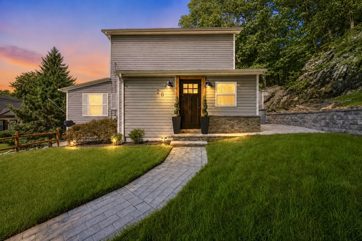 Twilight view of 26 Oak Street, Bloomingdale NJ — a renovated gray Colonial with warm-lit windows, a stone paver walkway across the front lawn, and a wooded rock hillside rising behind. Listed by BB Angjushev at Sotheby's International Realty.